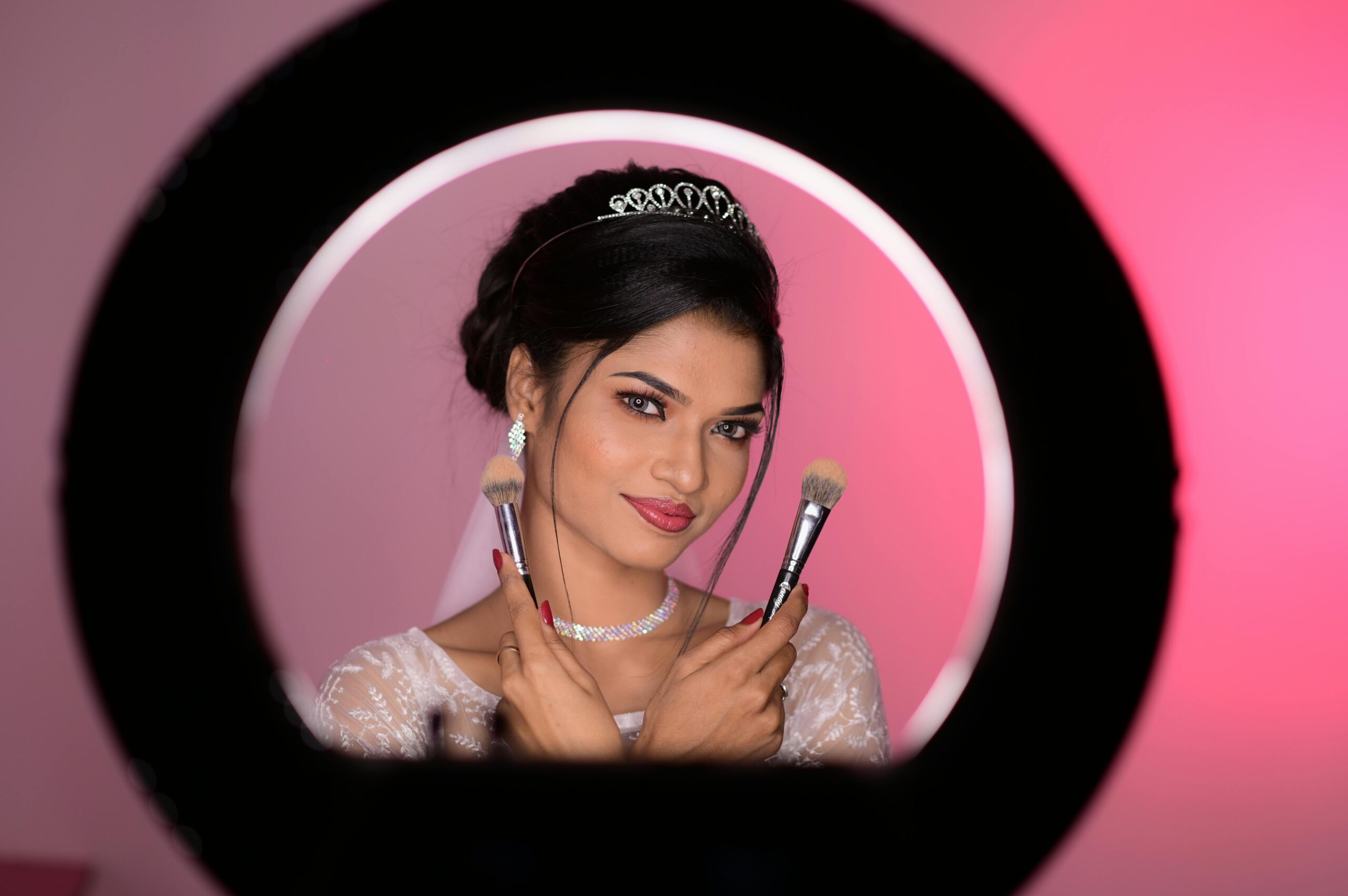 A beautiful bride poses with makeup brushes, framed by a ring light, in an elegant setting.